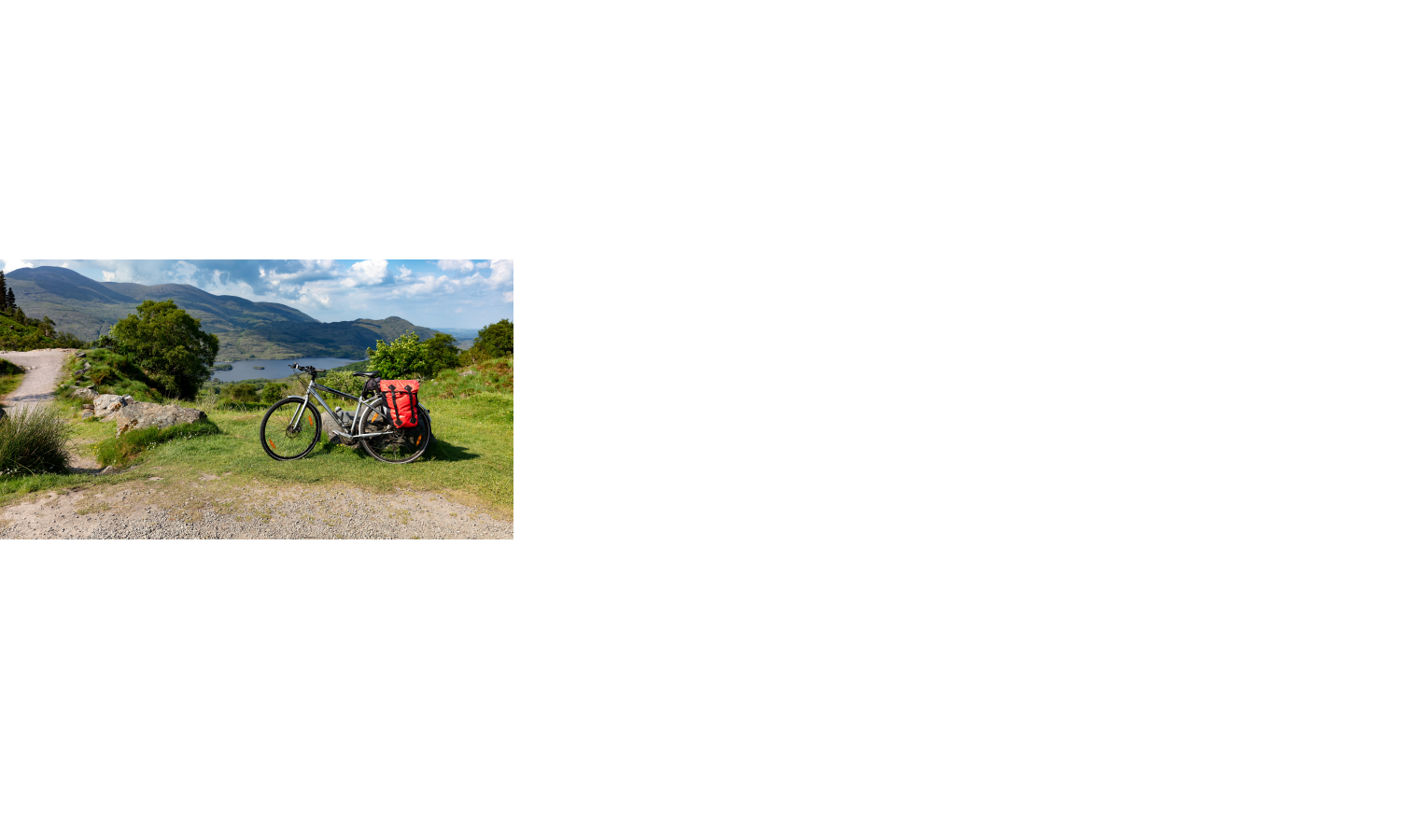 Fahrrad mit roter Tasche steht auf grünem Hügel, im Hintergrund See und Berge unter blauem Himmel.