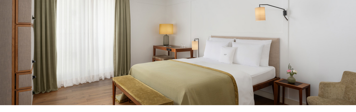 Gemütliches Hotelzimmer mit Doppelbett, grüner Tagesdecke, Holzbank, Schreibtisch und warmem Licht.