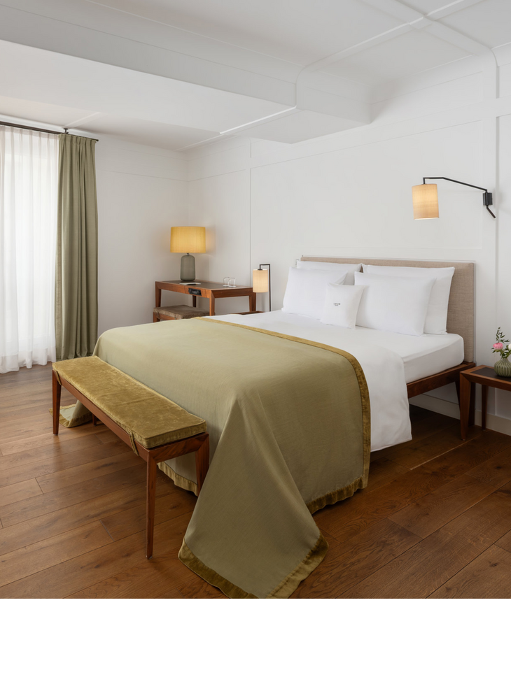 Gemütliches Hotelzimmer mit Doppelbett, grüner Tagesdecke, Holzbank, Schreibtisch und warmem Licht.