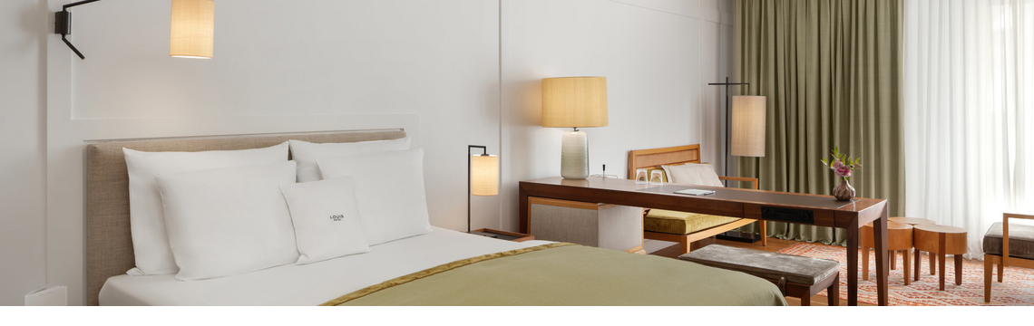Comfortable hotel room with double bed, desk, armchairs and large windows with curtains at the LOUIS Hotel Munich.