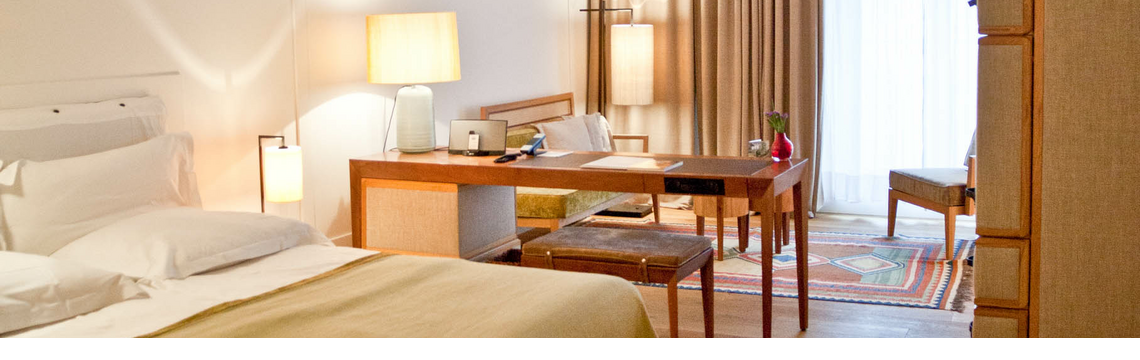 Comfortable hotel room with double bed, desk, armchairs and large windows with curtains at the LOUIS Hotel Munich.