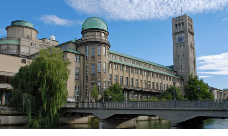 Historisches Gebäude mit grüner Kuppel und Turm am Fluss, daneben eine Brücke und Bäume, blauer Himmel im Hintergrund.