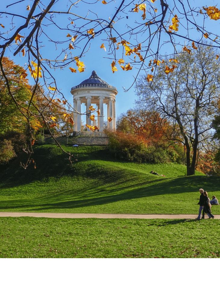 Temple in Munich's English Garden, surrounded by autumnal trees and green meadows, blue sky in the background.