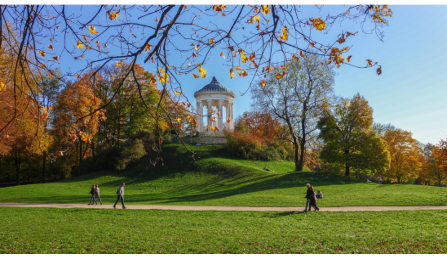 Der Monopteros im Englischen Garten mit Herbstbäumen und Spaziergängern auf grüner Wiese unter blauem Himmel.