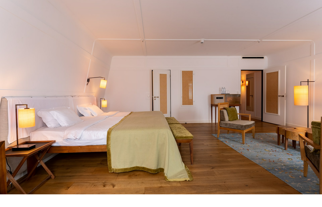 Bright, modern hotel room with large bed, wooden floor, stylish lamps and seating area in the LOUIS Hotel Munich.