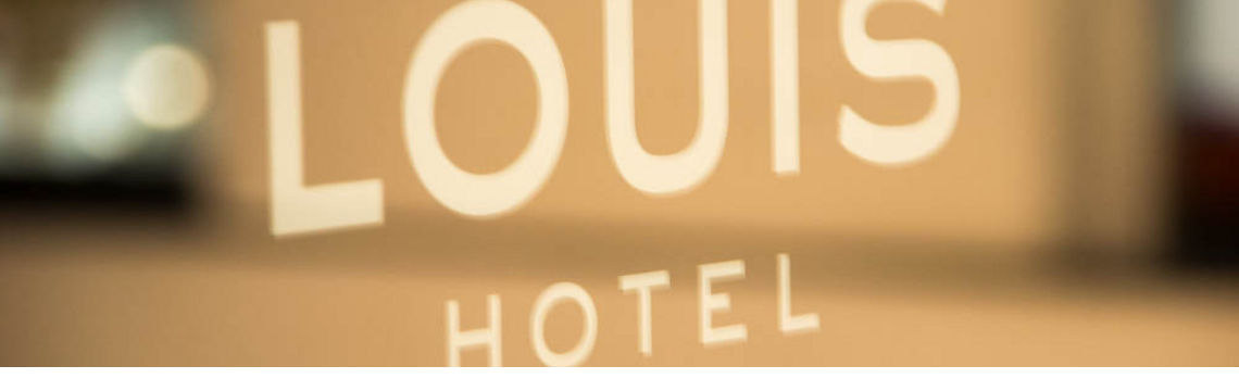 Aussenlogo an Glaswand LOUIS Hotel Muenchen Schriftzug "LOUIS HOTEL" auf Glas, unscharfer Hintergrund.