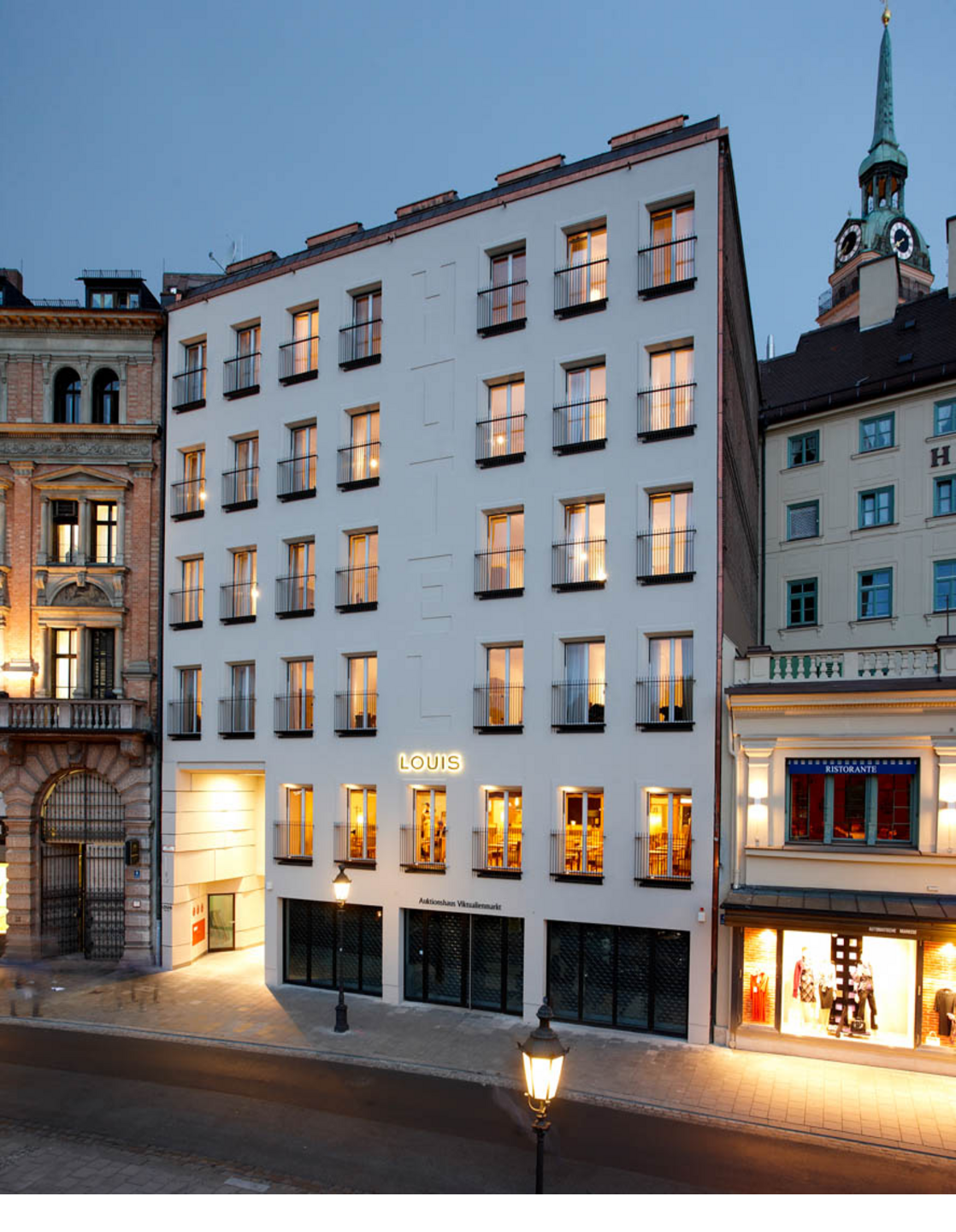 Aussenansicht nachts LOUIS Hotel Muenchen Außenansicht des LOUIS Hotels in München bei Dämmerung, mit beleuchteten Fenstern und umliegenden Gebäuden.