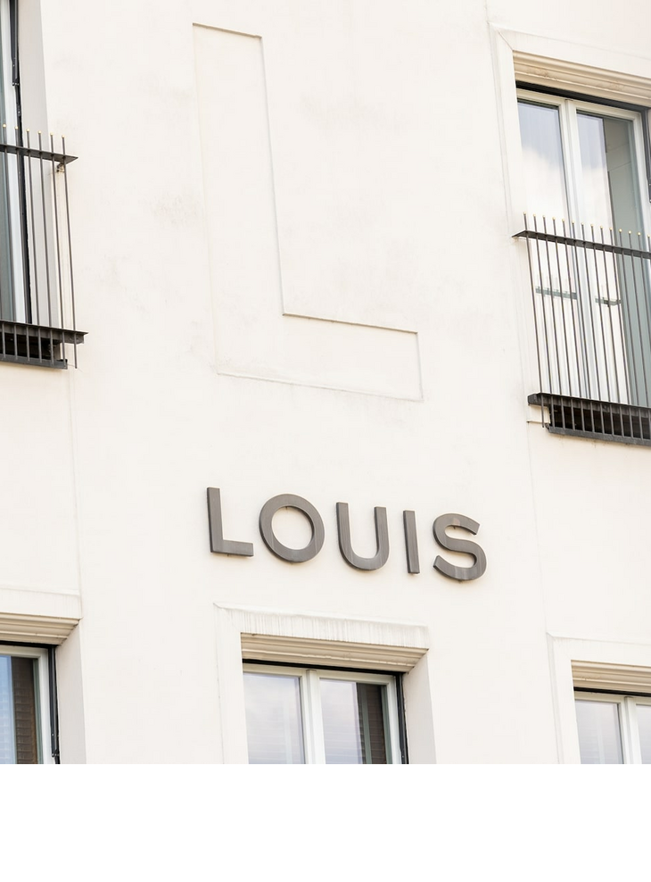 Exterior view LOUIS Hotel Munich logo Viktualienmarkt Façade of the LOUIS Hotel in Munich with large windows and balcony grilles.