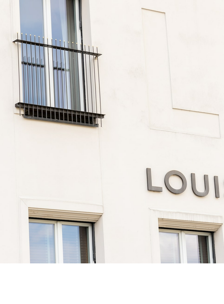 Aussenansicht LOUIS Hotel Muenchen Logo Viktualienmarkt Fassade des LOUIS Hotels in München mit großen Fenstern und Balkongittern.