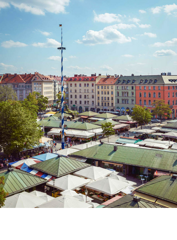 Blick auf den bunten Viktualienmarkt in München mit Maibaum, umgeben von historischen Gebäuden bei blauem Himmel.