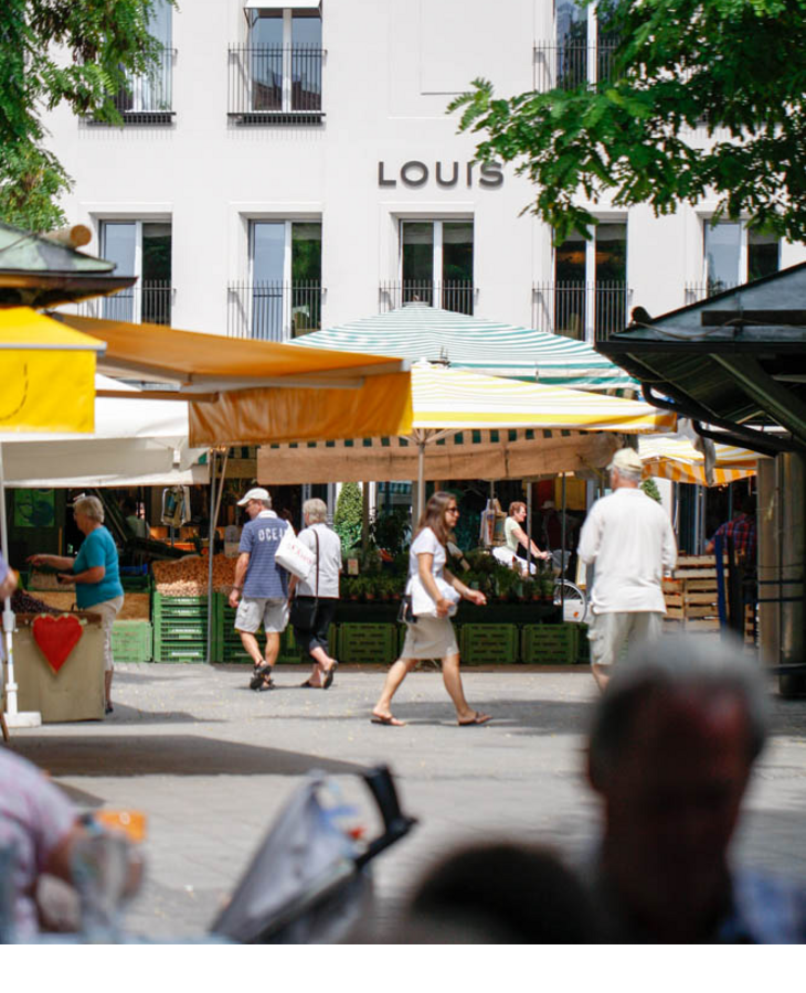 View of the LOUIS Hotel Munich with colorful market stall in the foreground and passers-by.
