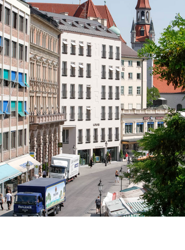 LOUIS Hotel in München, modernes Gebäude in urbaner Umgebung, mit Marktständen und Kirchturm im Hintergrund.