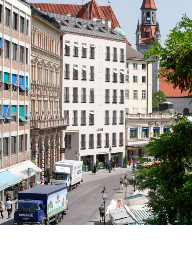 LOUIS Hotel in Munich, modern building in urban surroundings, with market stalls and church tower in the background.