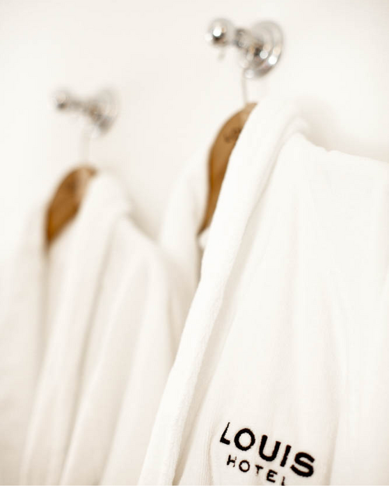 Bademantel im Badezimmer LOUIS Hotel Muenchen Zwei weiße Bademäntel mit LOUIS Hotel-Logo hängen an Haken an der Wand.
