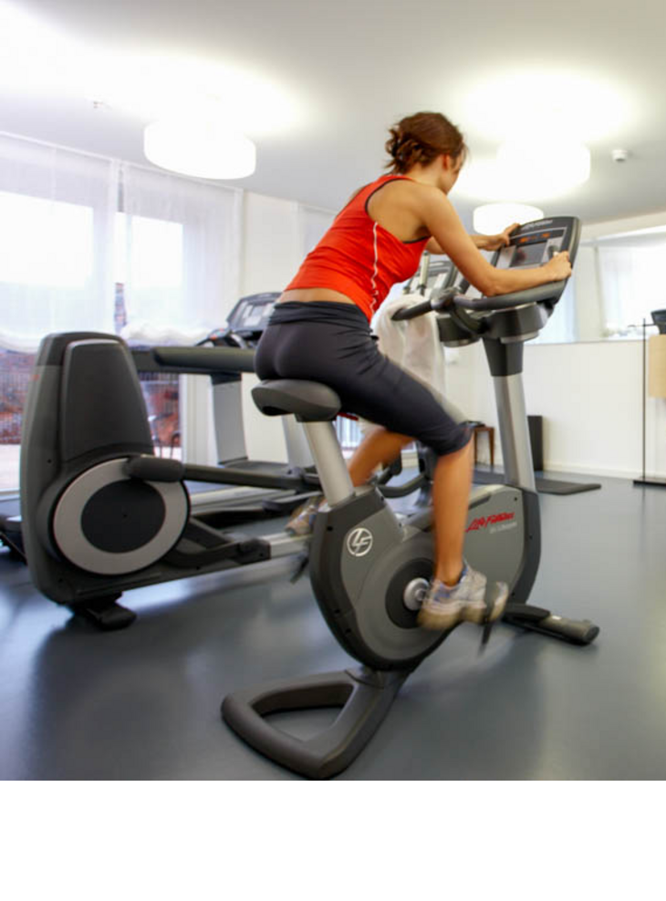 Frau trainiert auf einem Heimtrainer im Fitnessraum des LOUIS Hotels München, umgeben von Fitnessgeräten.