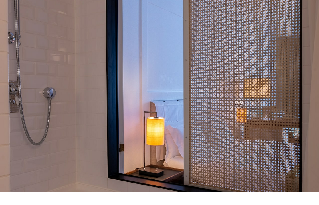 Hotel bathroom with bathtub, shower head and window to the bedroom with lamp and view of a bed in the LOUIS Hotel Munich.