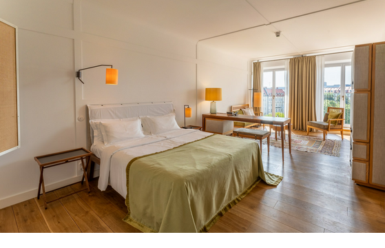 Bright hotel room with double bed, wooden floor, desk, seating area and large window with curtains.