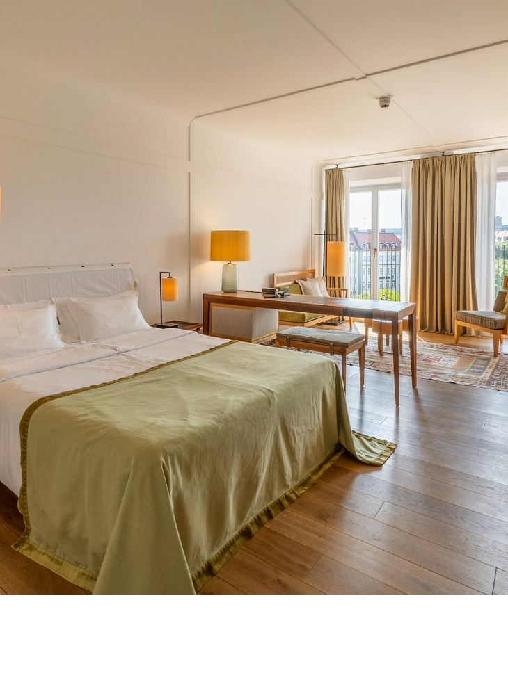 Helles Hotelzimmer mit Doppelbett, Holzboden, Schreibtisch, Sitzbereich und großem Fenster mit Vorhängen.