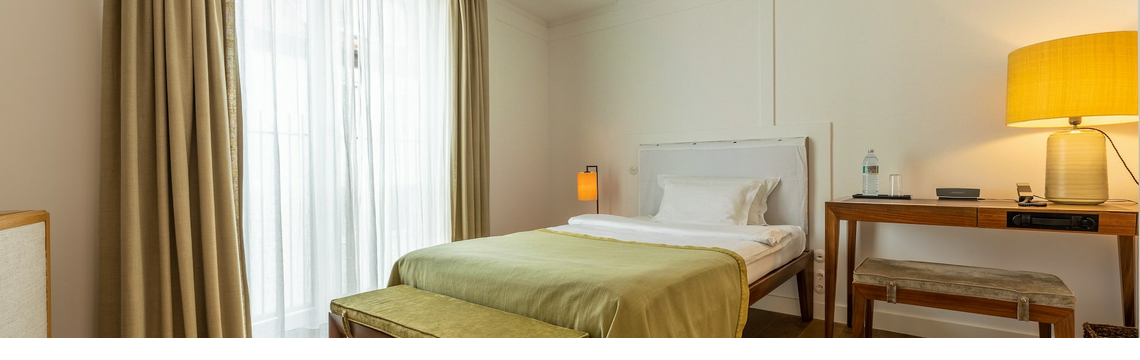 Bright hotel room with single bed, wooden floor, desk, lamp and large windows with curtains.