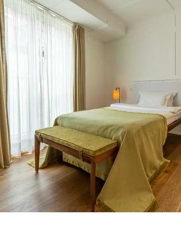 Bright hotel room with single bed, wooden floor, desk, lamp and large windows with curtains.