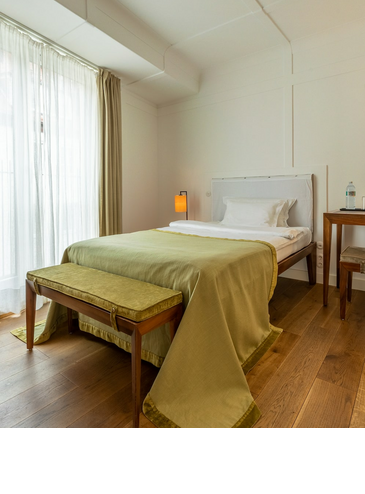 Helles Hotelzimmer mit Einzelbett, Holzboden, Schreibtisch, Lampe und großen Fenstern mit Vorhängen.