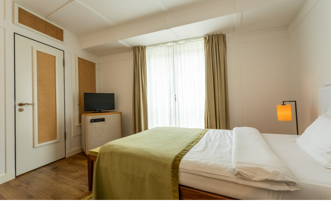 Bright hotel room with double bed, green bedspread, TV, lamp and large windows with curtains.