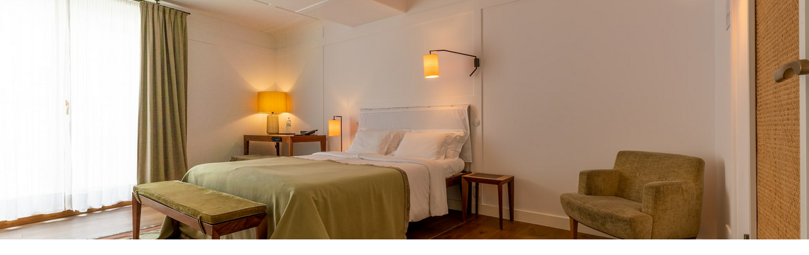 Bright hotel room with double bed, bedside tables, armchair, desk and lamp, wooden floor and curtains.