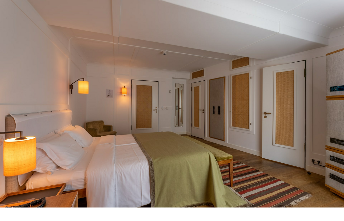 Bright hotel room with double bed, green bedspread, bedside lamps, carpet and armchair in the LOUIS Hotel Munich.