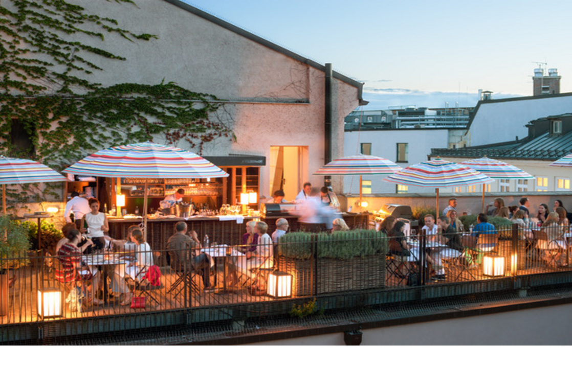 Roof terrace of the LOUIS Hotel in Munich with guests, colorful parasols and a cozy evening atmosphere.