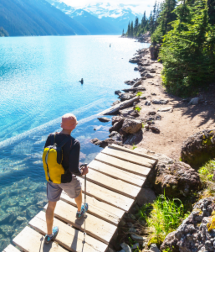 A man walks along a wooden footbridge by a turquoise mountain lake, surrounded by forest and mountains.