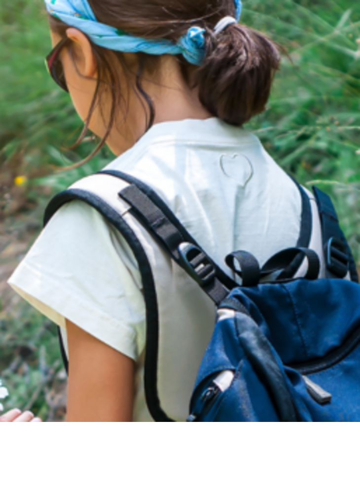 Two children with rucksacks are exploring a green meadow, one is holding a daisy flower.