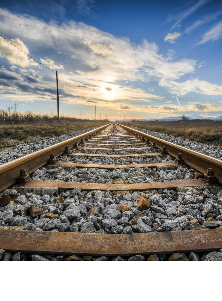 Mit der deutschen Bahn zu den ATLANTIC Hotels und ins LOUIS Hotel München reisen Gleise führen in den Sonnenuntergang, umgeben von Kies und Wiesen unter einem teils bewölkten Himmel.