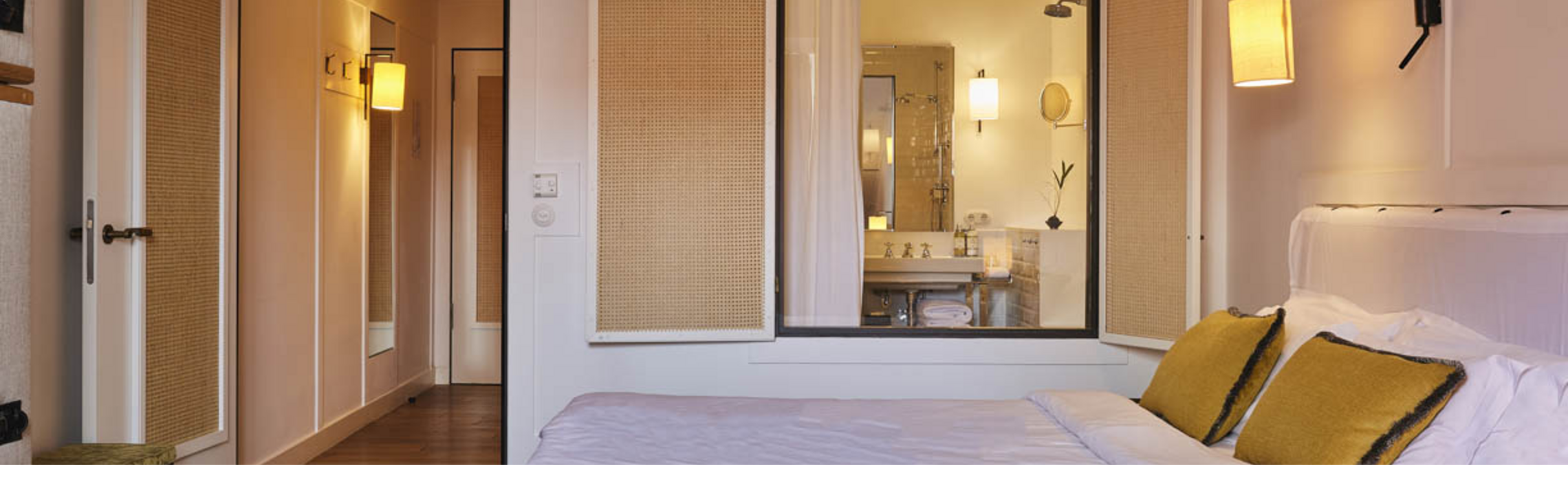 Modern hotel room with double bed, white sheets, yellow pillows, wooden floor and view of the bathroom through a window.