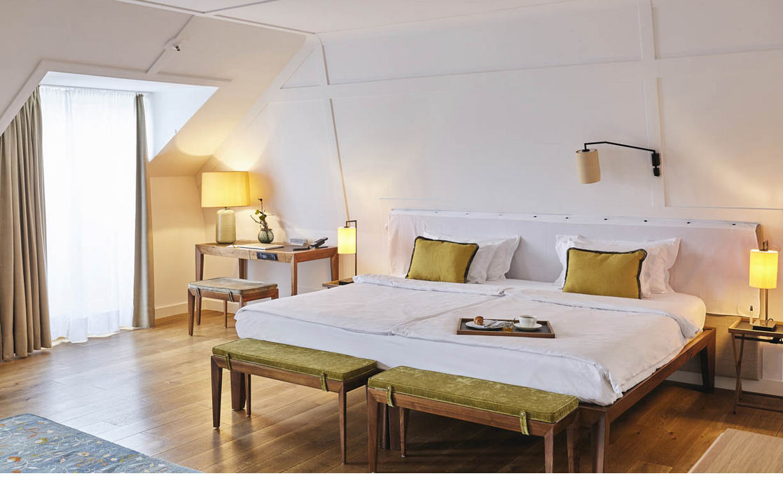 Doppelbett in der Suite im LOUIS Hotel Muenchen Helles Hotelzimmer mit großem Bett, Holzboden, Schreibtisch, zwei Hockern und einem Fenster mit Vorhängen.