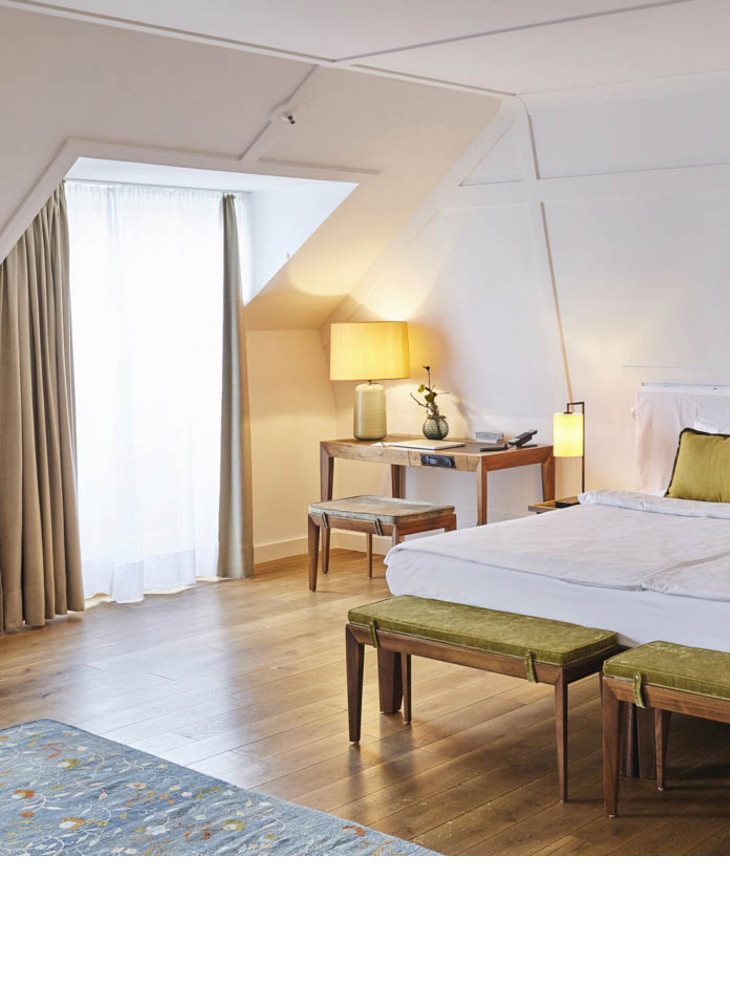 Helles Hotelzimmer mit großem Bett, Holzboden, Schreibtisch, zwei Hockern und einem Fenster mit Vorhängen.