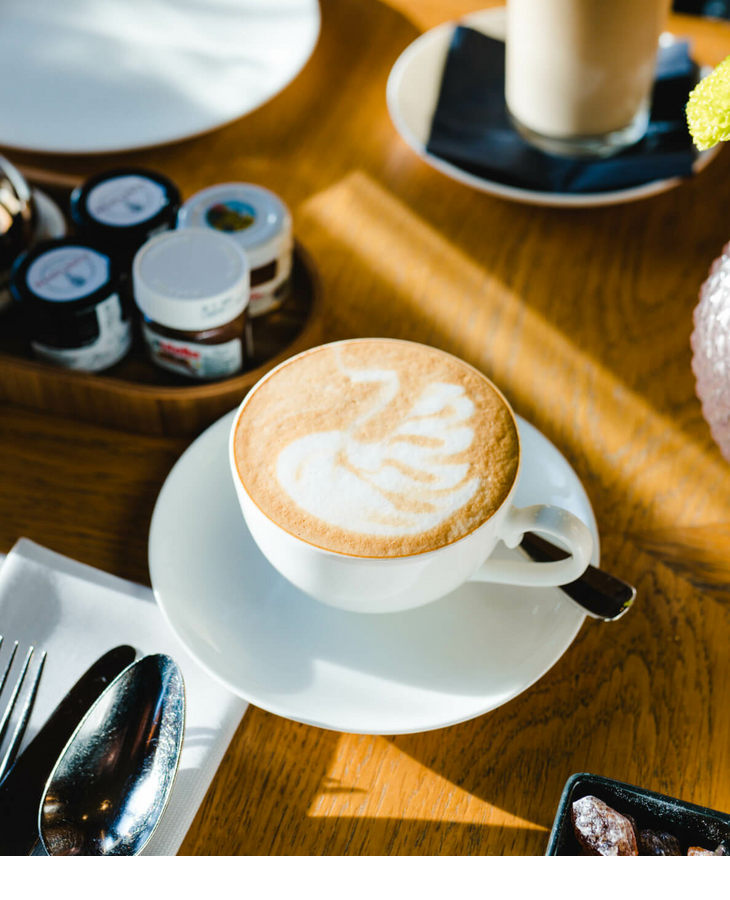 Tasse Cappuccino mit Latte Art auf Holztisch, umgeben von Marmeladengläsern und Besteck im LOUIS Hotel München.