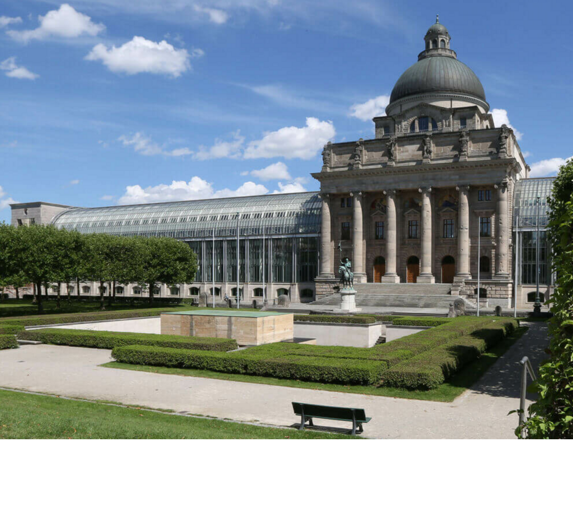 Historic building with dome and columns, surrounded by manicured gardens and hedges under a blue sky.