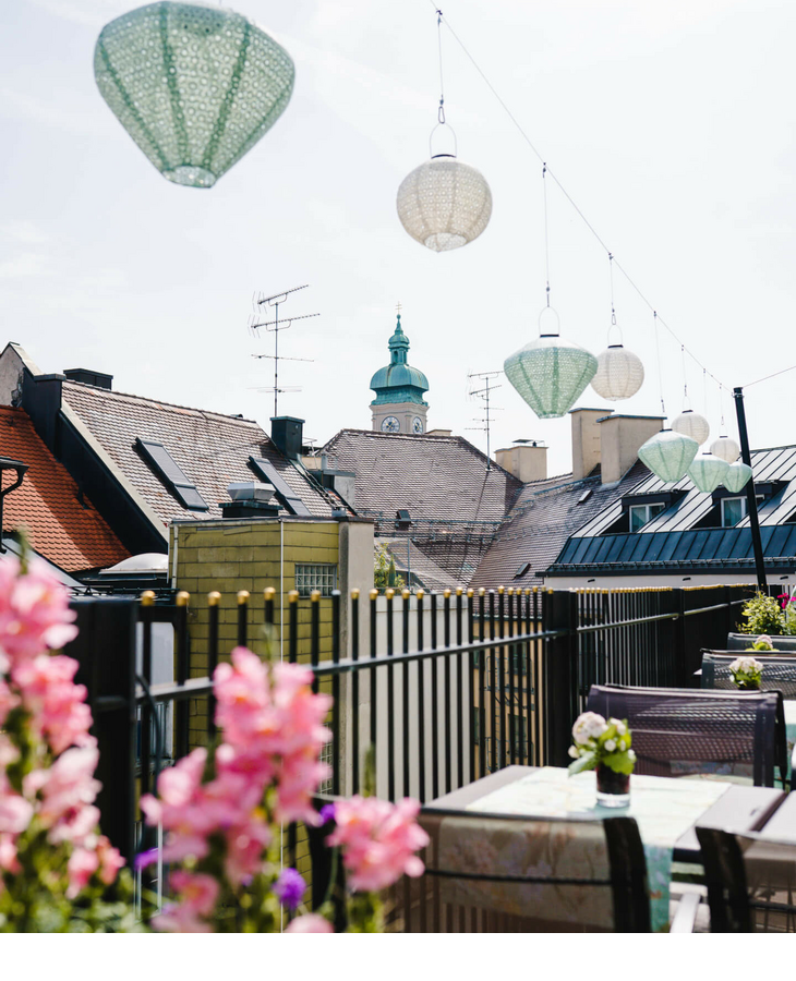 View from the hotel terrace with flowers, hanging lamps and church tower in the background.