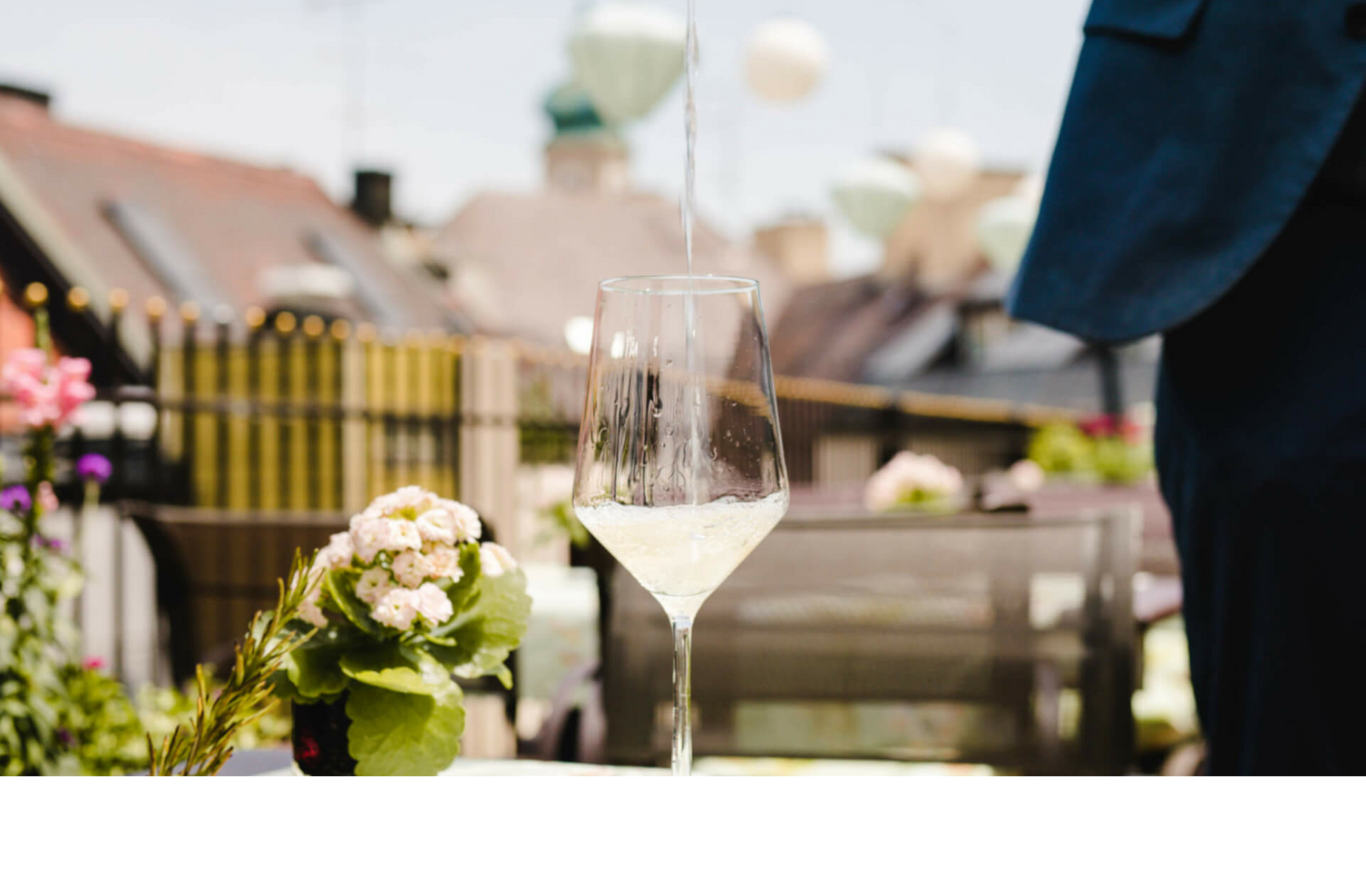Weißwein wird auf einer sonnigen Hotelterrasse in ein Glas eingeschenkt, mit Blumen und Stadtblick im Hintergrund.