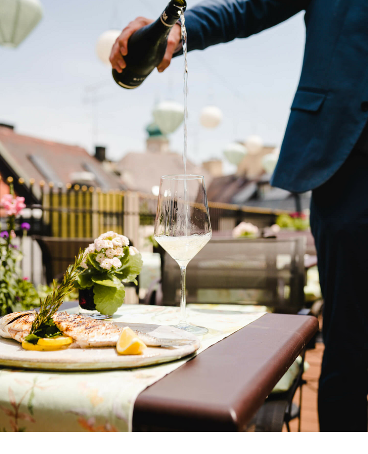 Person in Anzug schenkt Sekt auf sonniger Hotelterrasse ein, daneben ein Teller mit gegrilltem Fisch und Zitrone.