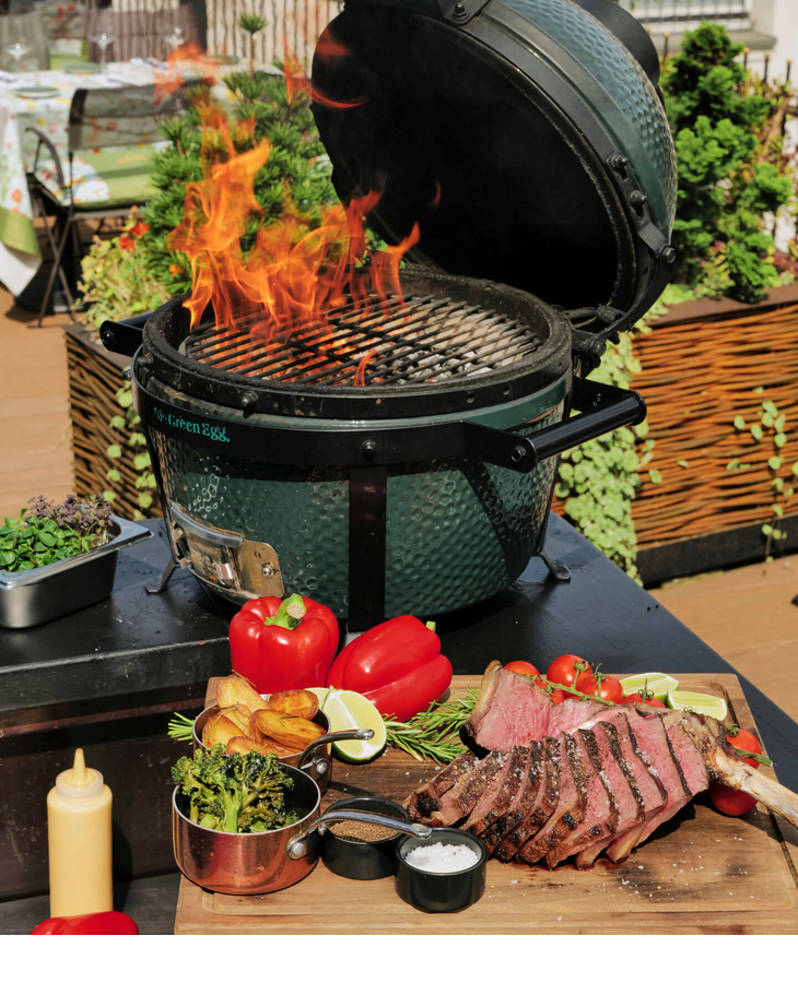 Grill mit offenen Flammen, davor ein Holzbrett mit gegrilltem Fleisch, Paprika und Beilagen auf einer Terrasse.