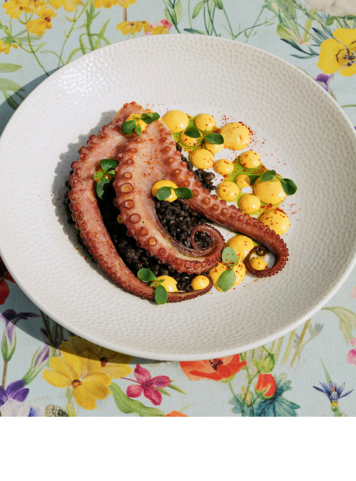 Grilled octopus on black rice with yellow sauce, served on a flowery table.
