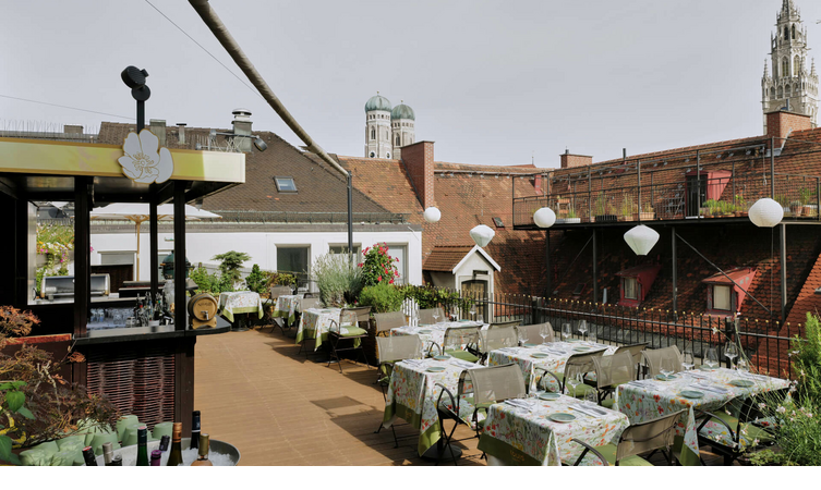 Roof terrace of the LOUIS Hotel Munich with covered tables, plants and a view of the towers of the Frauenkirche.