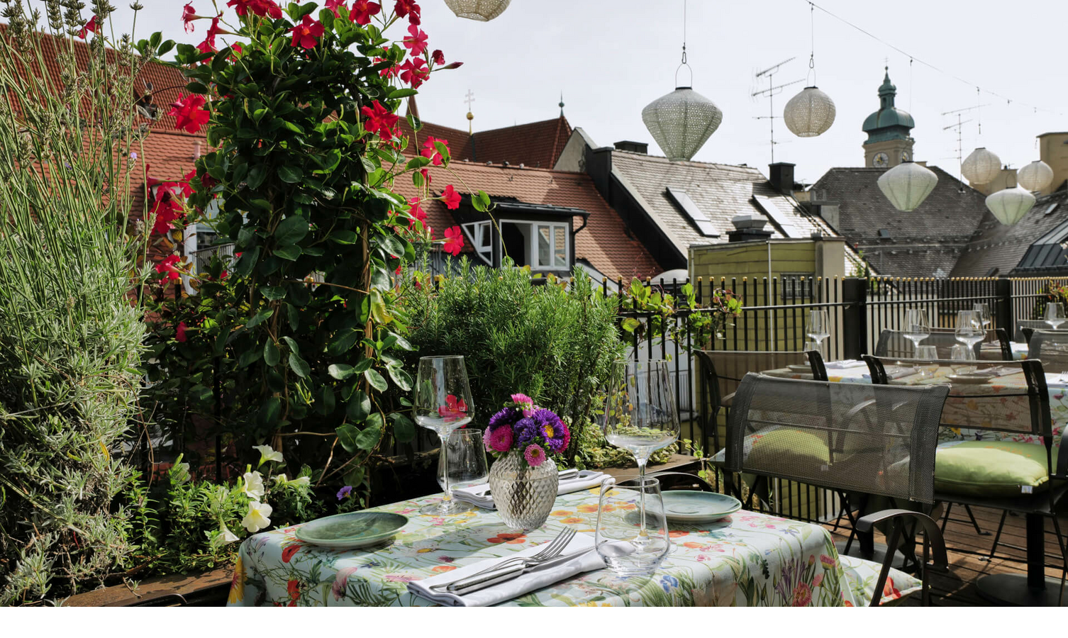 Roof terrace of the LOUIS Hotel with a table decorated with flowers, surrounded by plants and a view of Munich's rooftops.
