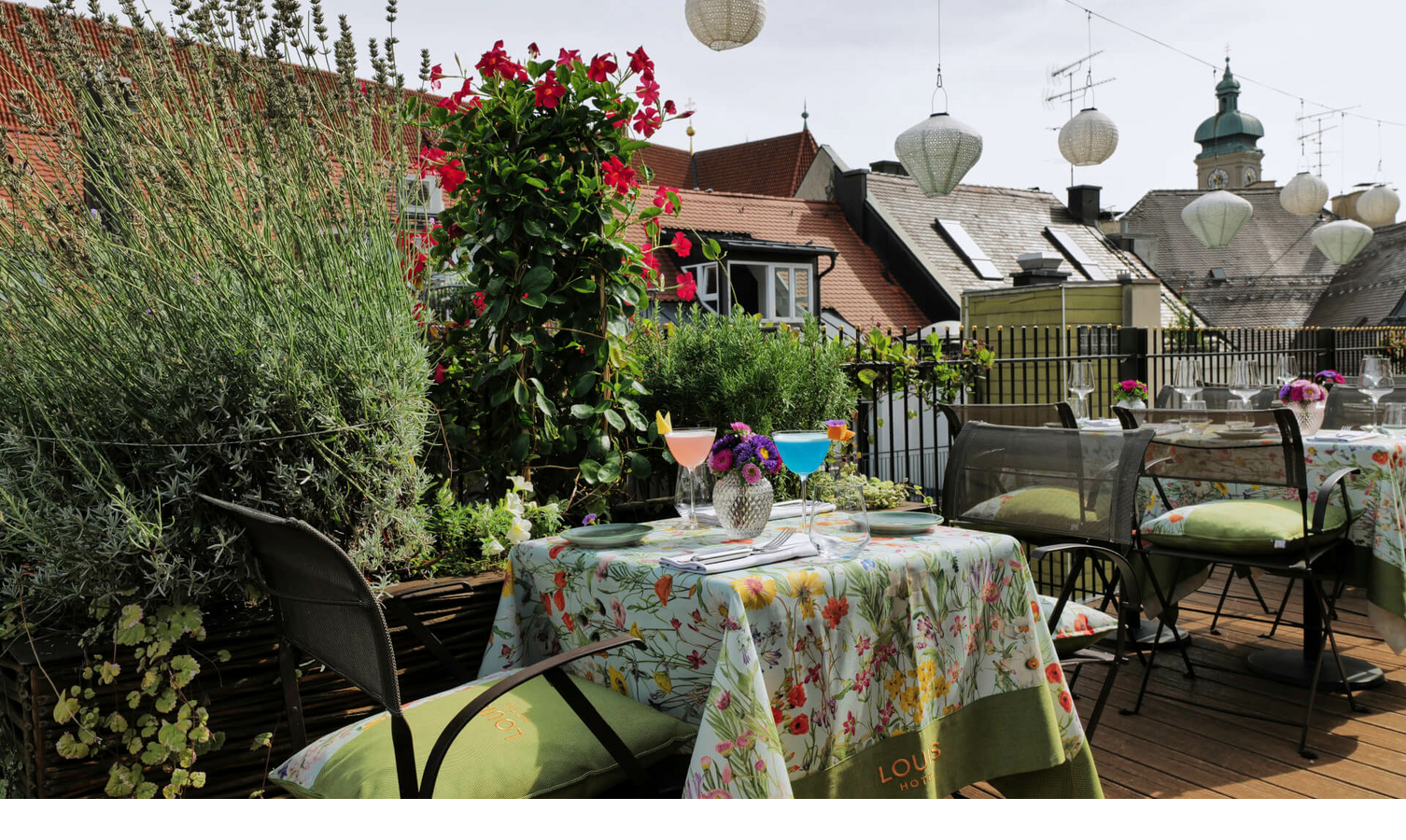 Dachterrasse des LOUIS Hotels in München mit blumengedecktem Tisch, Cocktails und Blick auf Dächer und Laternen.