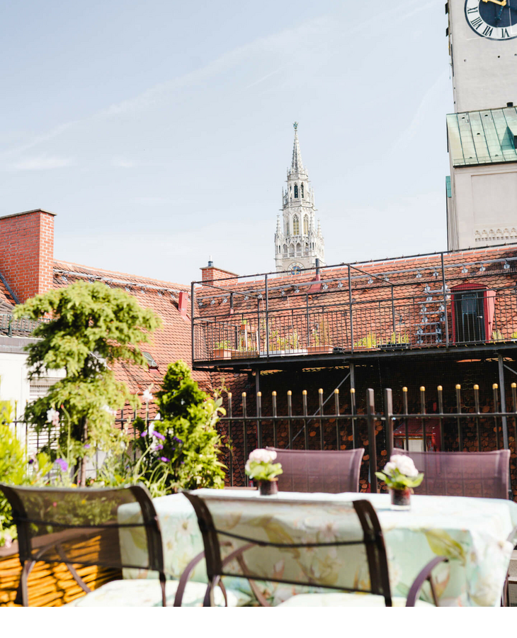 The Roof Terrace – Dachterrasse mit herrlichem Blick | LOUIS Hotel München Dachterrasse mit Sitzbereich und Pflanzen, Blick auf Münchens Kirchturm bei blauem Himmel.