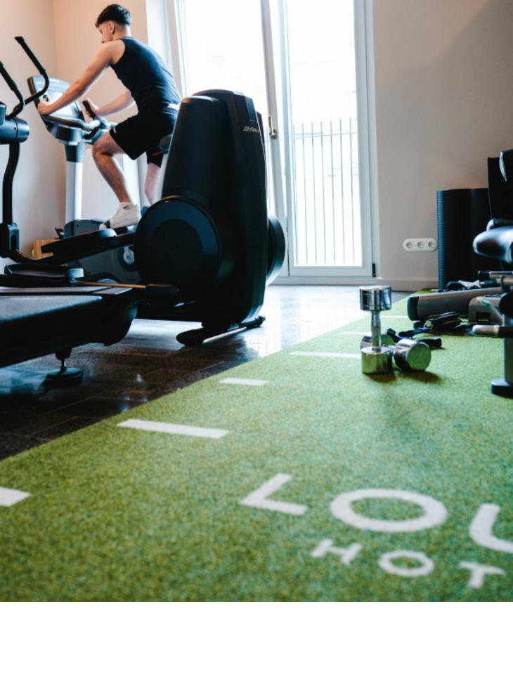 Person trainiert im Fitnessraum des LOUIS Hotels München auf einem Crosstrainer, Hanteln auf grünem Teppich.