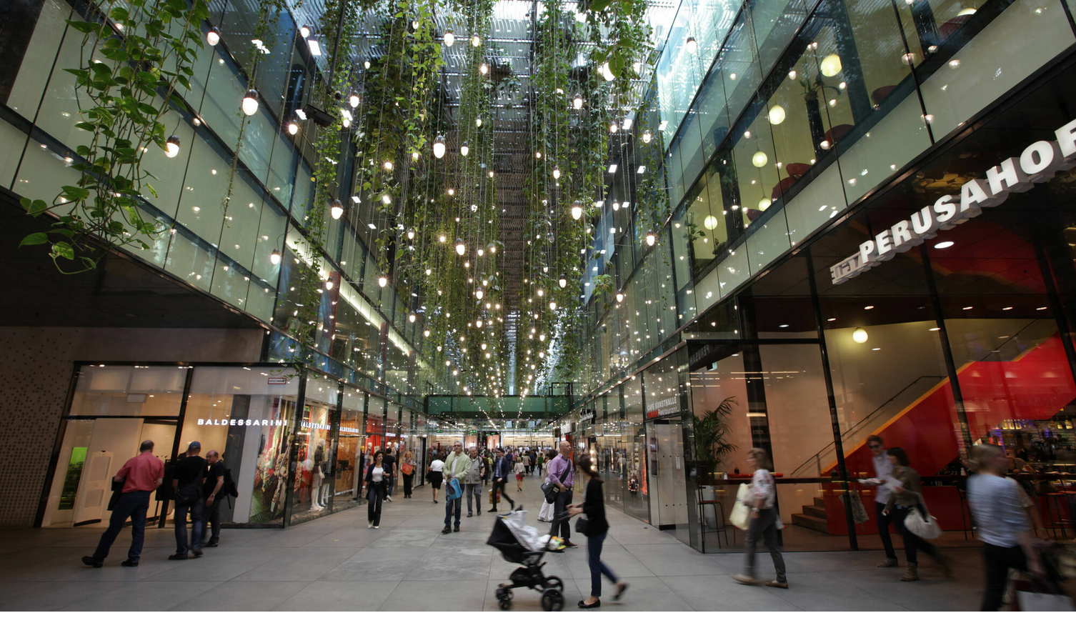 Shopping arcade with glass roof, hanging plants and lights, surrounded by stores and people out for a stroll.