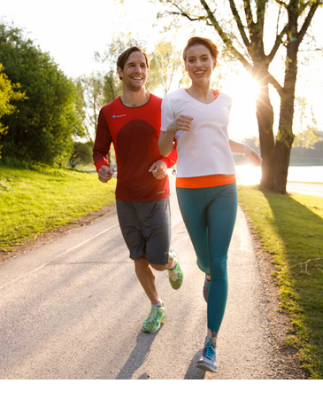Gemeinsam Joggen in München Zwei Personen joggen lachend auf einem sonnigen Parkweg, umgeben von Bäumen und Grünflächen.