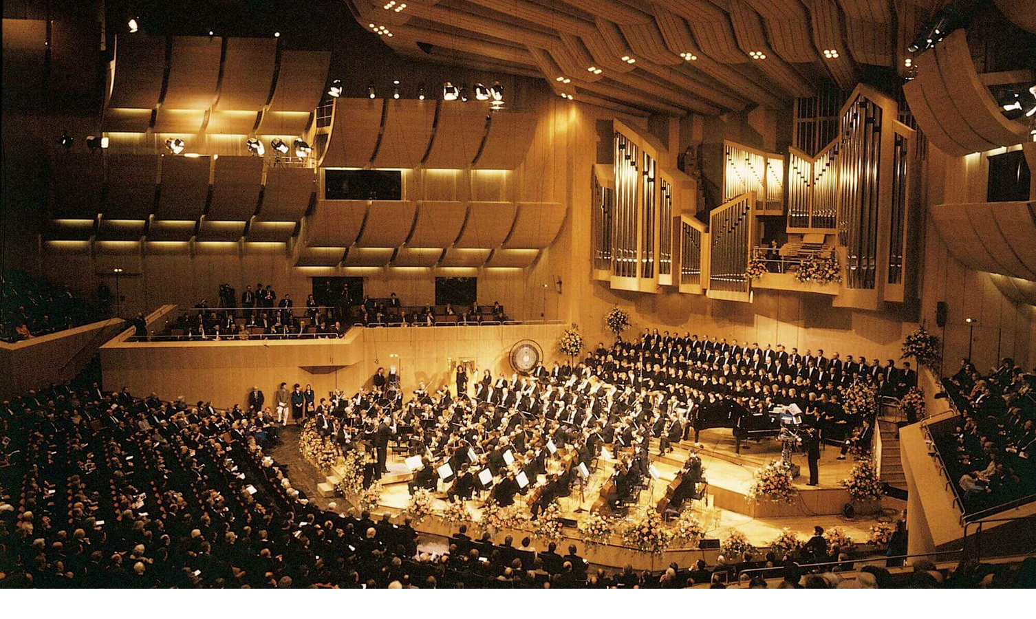 Ein Konzertsaal mit Orchester und Chor auf der Bühne, umgeben von Publikum in einem modernen, holzverkleideten Raum.